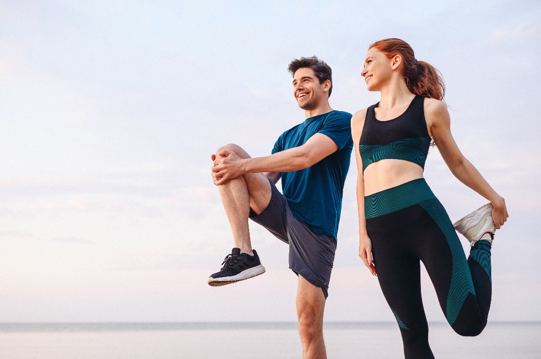 Ein junges Paar, zwei Freunde, sportlich, Sportlerin, Sportler, Frau, Mann, in Sportbekleidung, Aufwärmen, Training, Dehnübungen, auf Sand, Meer, Ozean, Strand, im Freien, Joggen, am Meer, an einem Sommertag, morgens.