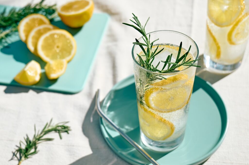 Ein erfrischendes Sommergetränk – Limonade oder ein alkoholischer Cocktail mit Eis, Rosmarin und Zitronenscheiben – auf dem Tisch im Garten. Ein frisches, gesundes, kaltes Zitronengetränk. Wasser mit Zitrone.